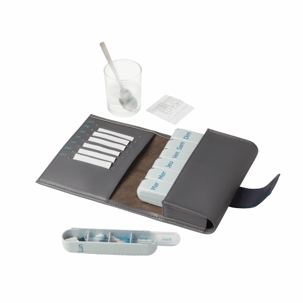 Pilbox Cardio gris ouvert avec six modules à l'intérieur, un module posé devant avec des médicaments dedans et un verre avec une cuillère au dernier plan avec un médicament soluble à côté